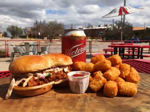 A crispy chicken sandwich with coleslaw, a side of tater tots, and a can of beer on a picnic table outdoors.