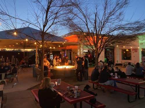 A lively outdoor gathering with people around a fire pit, string lights, and trees, creating a cozy evening atmosphere.