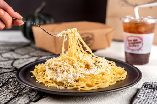 A fork twirls spaghetti topped with cheese on a black plate, with a drink and takeout box in the background.