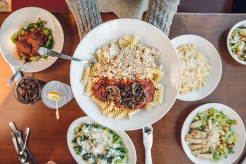 A person holds a plate of pasta with marinara sauce and mushrooms, surrounded by various dishes on a wooden table.