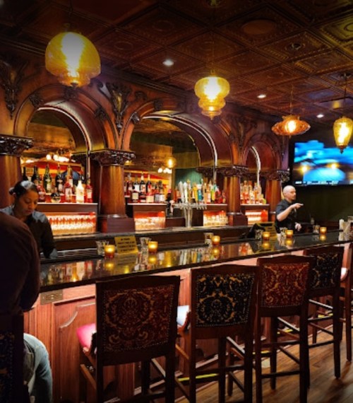 A stylish bar with ornate wooden decor, glowing lights, and a bartender serving drinks.
