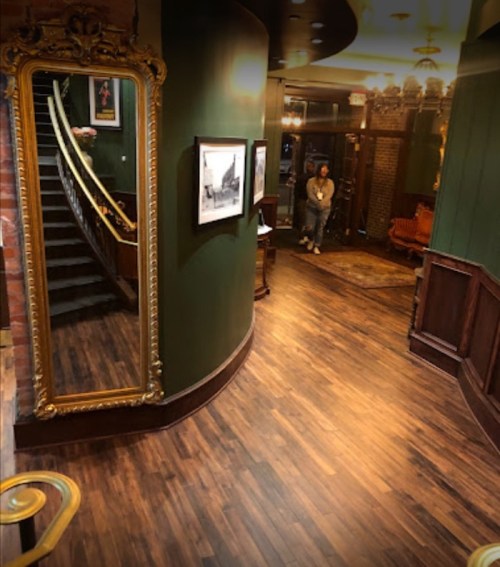 A dimly lit hallway with a large mirror, staircase, and vintage decor, featuring a person standing in the background.