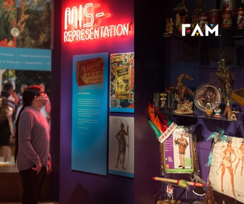 A visitor observes an exhibit titled "MIS-REPRESENTATION" featuring artifacts and neon signage in a museum setting.