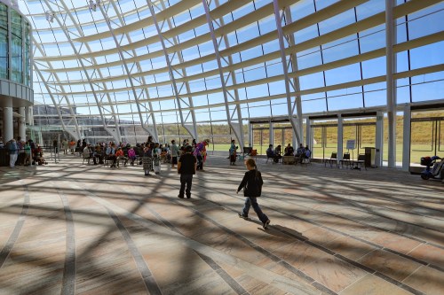 A spacious, modern interior with large windows, people seated and walking on a patterned floor, bathed in natural light.