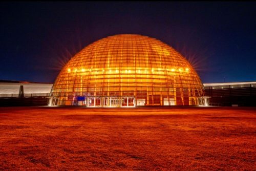 A large, illuminated glass dome structure at night, surrounded by a grassy area.