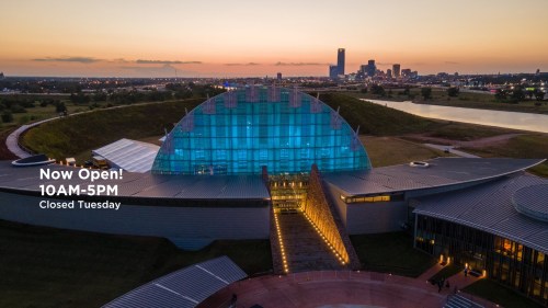 Aerial view of a modern building with a glass dome, surrounded by greenery and water, with a city skyline in the background.