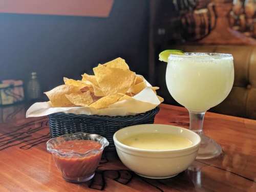 A basket of tortilla chips, a bowl of salsa, a bowl of queso, and a margarita with a lime wedge on a wooden table.