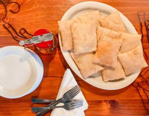 A plate of golden-brown pastries sits on a wooden table, accompanied by utensils and a container of seasoning.