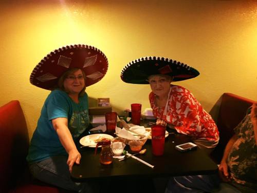 Two women wearing large sombreros sit at a table with food and drinks, smiling at the camera in a casual restaurant setting.
