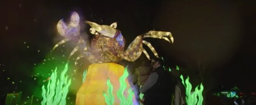 A large illuminated crab sculpture surrounded by colorful lights and green seaweed, with a person standing nearby.