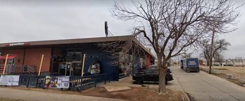 A building with a colorful facade, parked cars, and a bare tree in front, on a cloudy day.