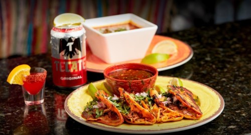 A plate of tacos with toppings, a bowl of soup, a can of beer, and a shot glass with a lemon slice.