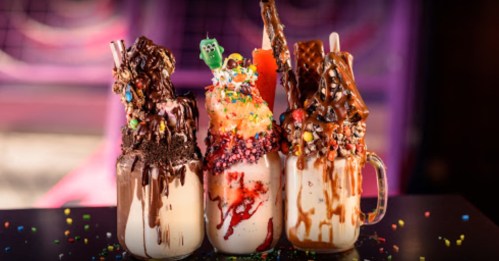 Three extravagant milkshakes topped with colorful candies, whipped cream, and chocolate drizzles, set against a vibrant background.