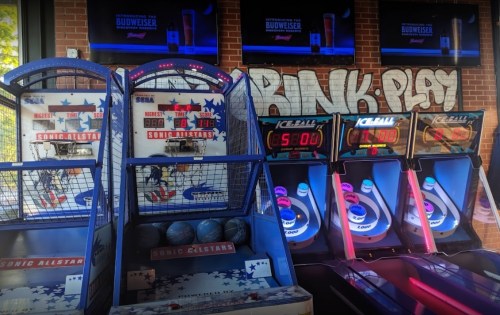 Two arcade games, a basketball shooter and an ice hockey game, with a "BANK PLAY" graffiti in the background.