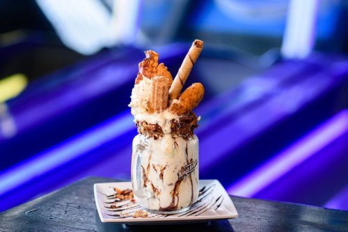 A decadent dessert in a jar, featuring ice cream, whipped cream, and various toppings, set against a colorful background.