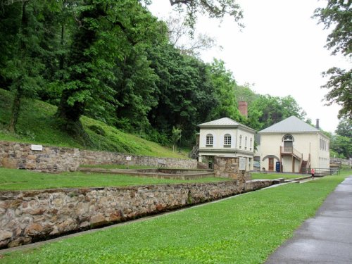 Historic building beside a stone canal, surrounded by lush greenery and trees on a cloudy day.