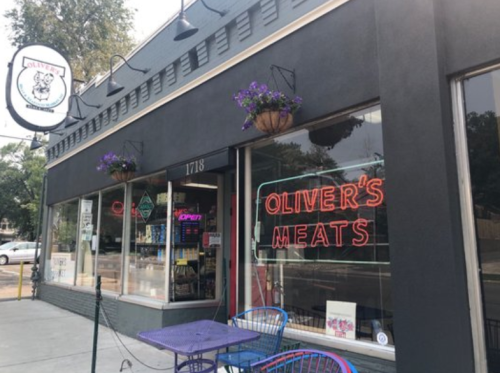 Exterior of Oliver's Meats shop with neon sign, potted flowers, and outdoor seating.