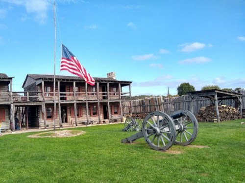 Historic fort with an American flag, cannons, and wooden buildings under a clear blue sky.