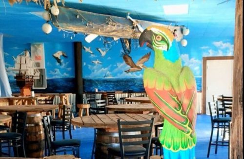 A colorful parrot statue stands in a vibrant restaurant with nautical decor and wooden tables.