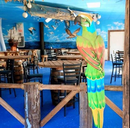 Colorful parrot statue stands near wooden tables and chairs in a beach-themed restaurant with a blue mural.
