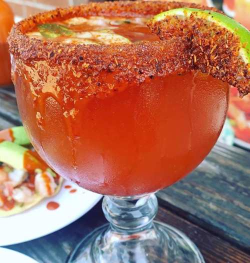 A large, rimmed cocktail glass filled with a spicy drink, garnished with a lime slice, next to a plate of shrimp.