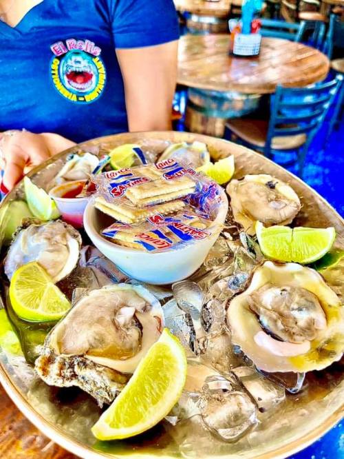 A person holds a platter of fresh oysters with lemon wedges and sauces on a table in a restaurant.