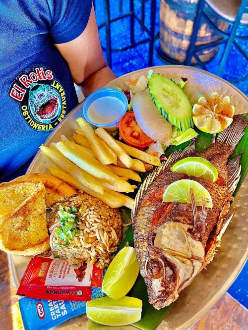 A plate of grilled fish with lime, fries, rice, salad, and sauces, served on a banana leaf.
