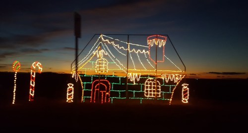 A brightly lit gingerbread house decoration with candy canes against a sunset sky.