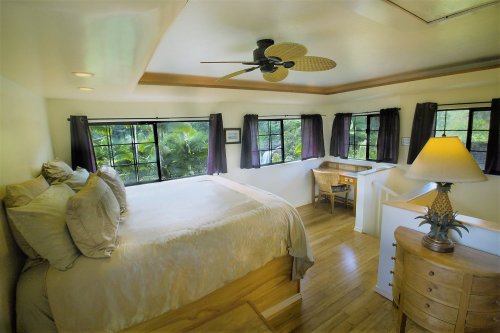 Cozy bedroom with a large bed, wooden floors, a ceiling fan, and windows overlooking greenery.