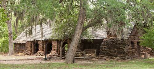 A rustic stone building surrounded by trees and greenery, featuring a sloped roof and moss hanging from branches.