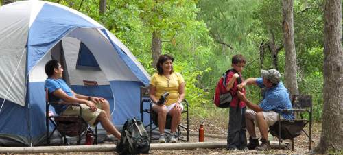 A family enjoys camping in the woods, sitting by a tent and interacting with each other.