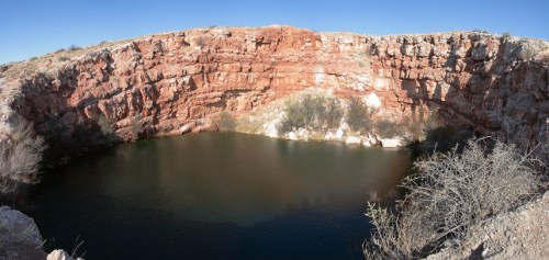 A panoramic view of a deep, rocky pit filled with dark water, surrounded by dry vegetation and clear blue sky.