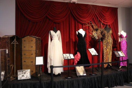 A display of elegant dresses on mannequins against a red backdrop, with vintage photos and a decorative wardrobe.