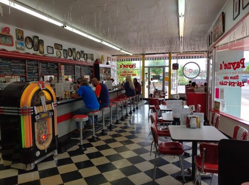 A retro diner interior with checkered floors, red booths, a jukebox, and customers seated at the counter.