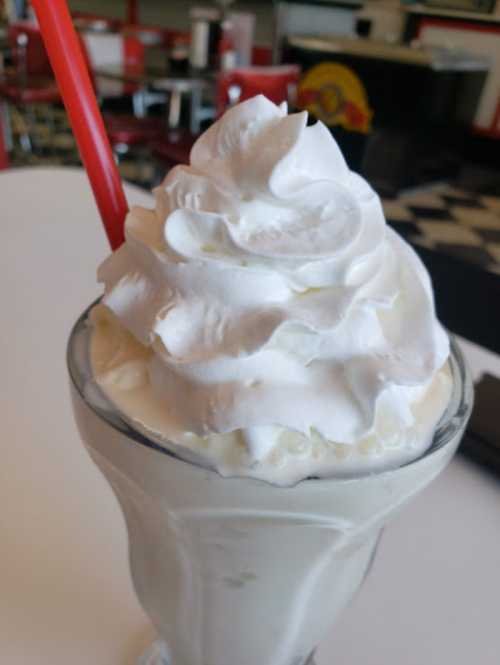 A tall glass of vanilla milkshake topped with a generous swirl of whipped cream and a red straw.