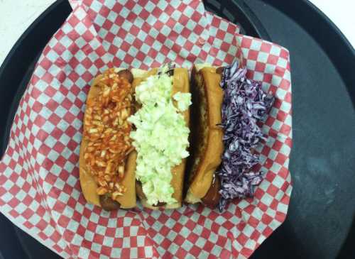 Three hot dogs on a red and white checkered paper, topped with chili, coleslaw, and purple cabbage, on a black tray.