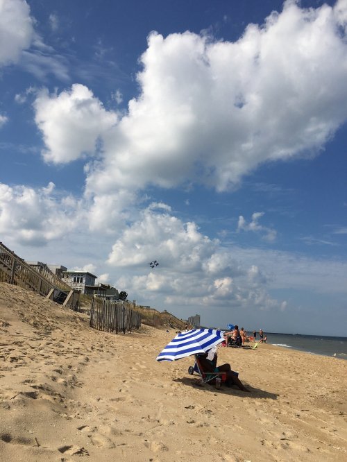A beach scene with people relaxing under umbrellas, sand dunes, and fluffy clouds in a blue sky.