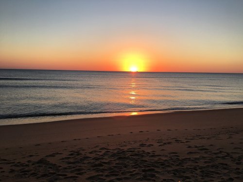 A serene beach at sunset, with vibrant orange and purple hues reflecting on calm water and footprints in the sand.