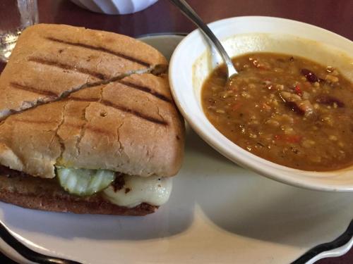 A grilled sandwich with pickles and melted cheese next to a bowl of hearty soup with vegetables and beans.
