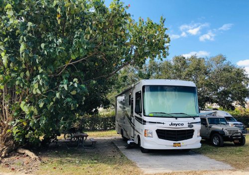 A white Jayco RV parked under a large tree, with a gray Jeep nearby and a picnic table in the background.