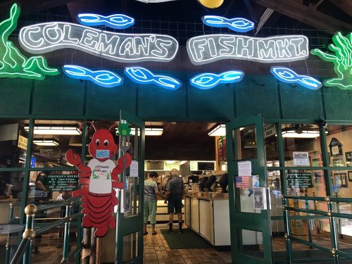 Entrance to Coleman's Fish Market, featuring a lobster mascot wearing a mask and neon signage. Customers inside.