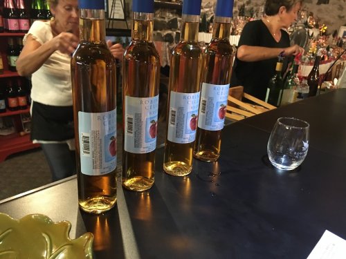 Four bottles of wine with blue caps on a bar, with a glass and people in the background.