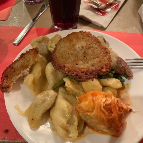 A plate of assorted dumplings, a crispy potato pancake, and a piece of baked meat, with a drink in the background.