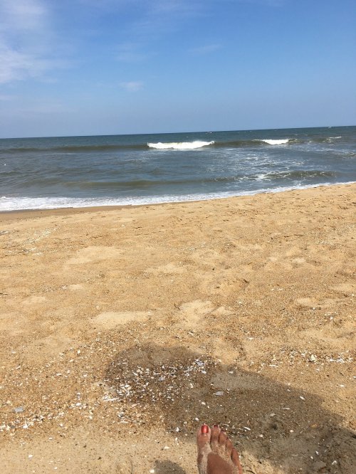 A sandy beach with gentle waves and a clear blue sky, featuring a close-up of a foot with red nail polish.