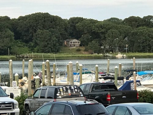 A view of a marina with boats docked, surrounded by trees and a house on a hill in the background.