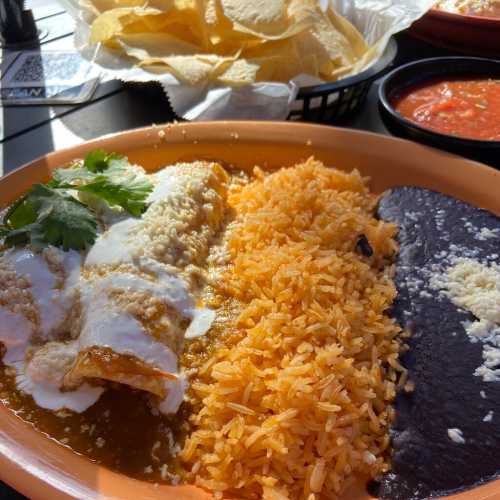 A plate of enchiladas topped with cheese and sauce, served with rice and black beans, alongside tortilla chips and salsa.