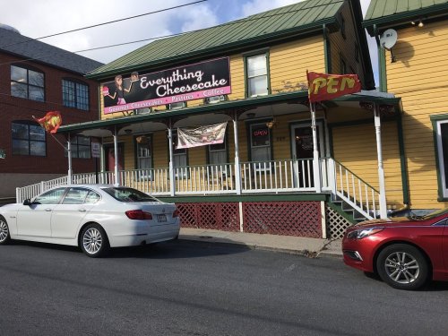 A yellow building with a sign reading "Everything Cheesecake," featuring flags and parked cars in front.