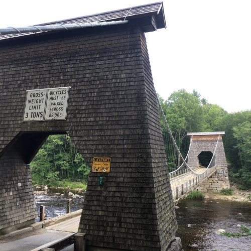A wooden suspension bridge spans a river, with a sign indicating a weight limit and bicycle restrictions.