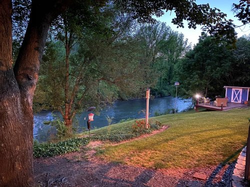 A serene riverside scene at dusk, featuring trees, a grassy area, and a cozy shed by the water.