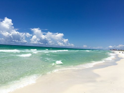 A serene beach scene with gentle waves, soft white sand, and a clear blue sky dotted with fluffy clouds.